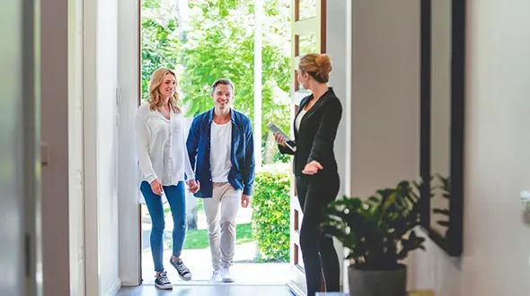 A couple walks through a hallway as a real estate agent holds a phone, discussing property details.