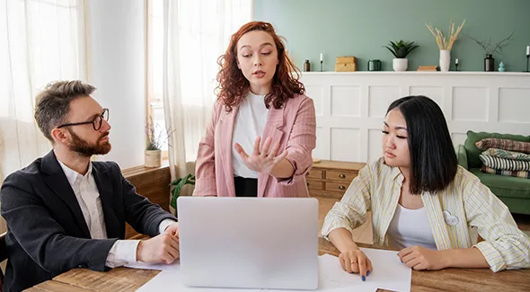 A group of three people engages around a table with a laptop, focusing on strategies for expanding real estate options.
