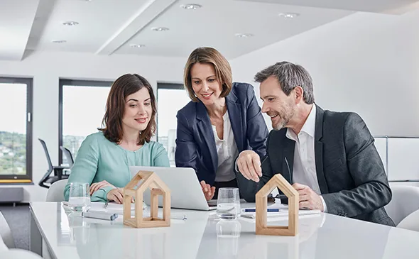 Three people sit around a table with a laptop and a house model, strategizing about local real estate opportunities.